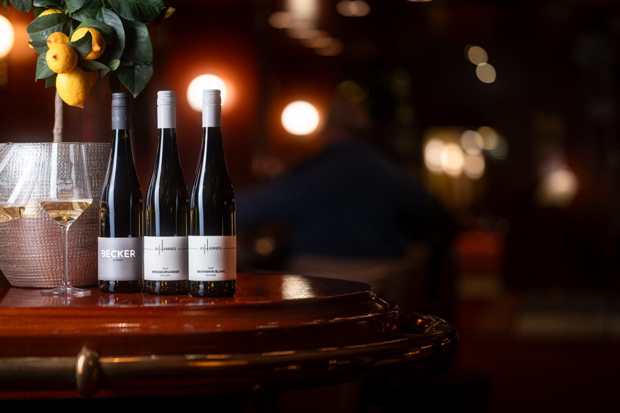 Drei Weinflaschen und ein Glas auf einem Bartresen, dahinter warme Lichter