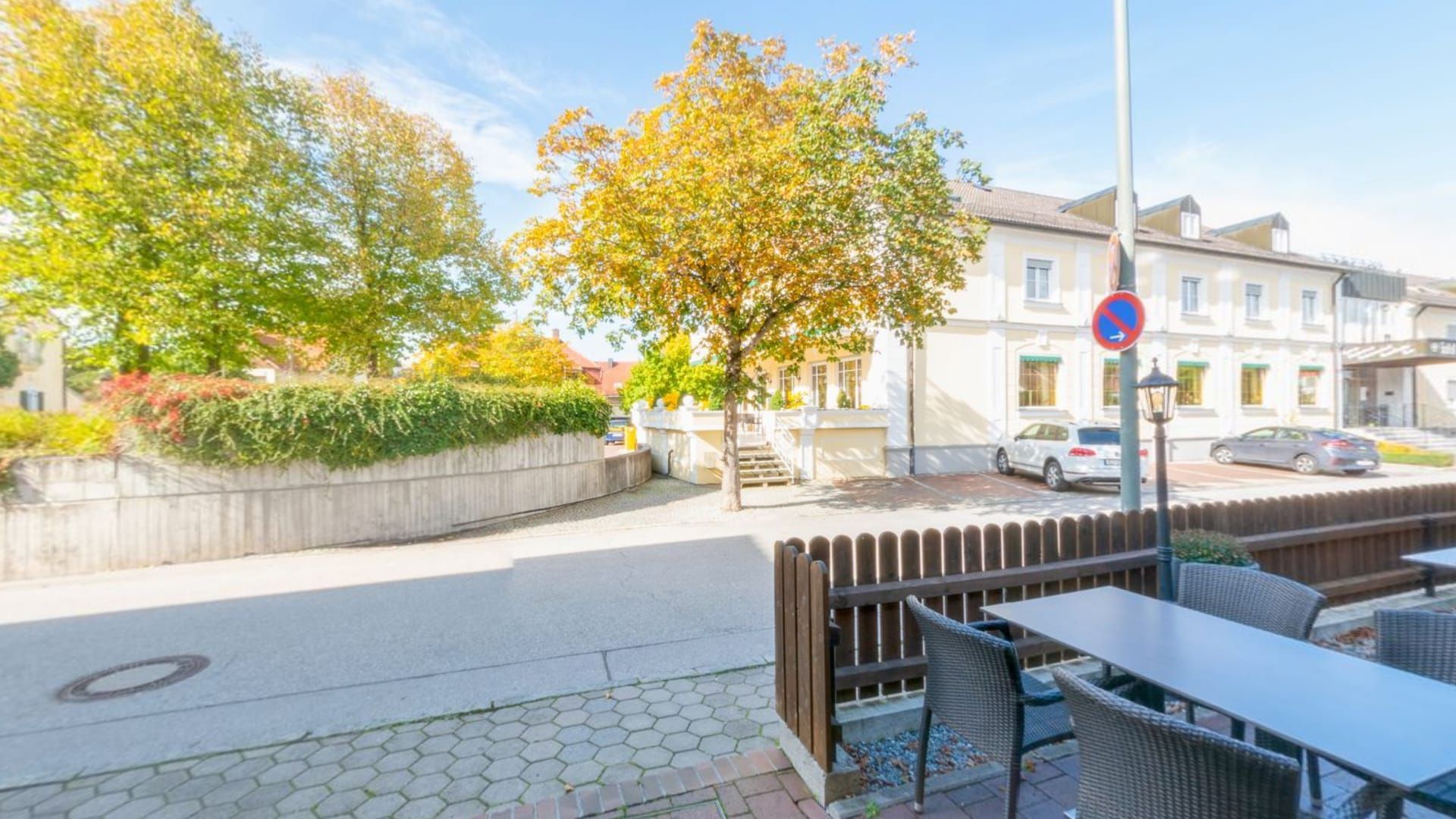 Terrasse vor dem Eingang mit Tischen, dahinter Straße und Herbstbäume